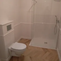 Nowoczesna łazienka z białą armaturą, kabiną prysznicową typu walk-in i podłogą z parkietu ułożonego we wzór jodełki.