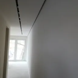 Korytarz w trakcie wykańczania, surowa ściana po gładzi, widoczne listwy oświetleniowe w suficie, okno z widokiem na zewnątrz.