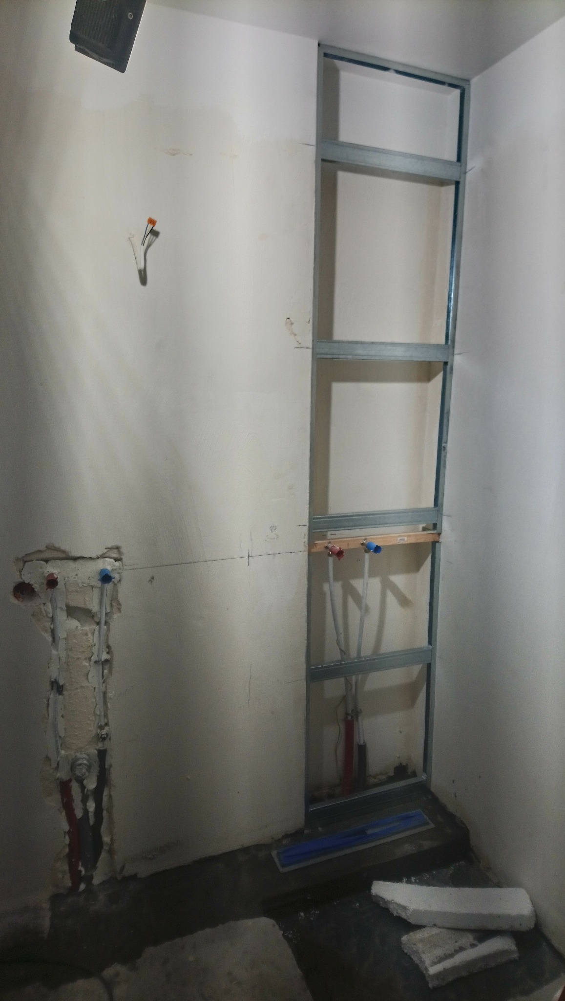 Stelaż z profili metalowych przy ścianie, przygotowane rury instalacyjne, fragment ściany z odkrytymi rurami, niebieska poziomica na podłodze, zaprawa murarska.