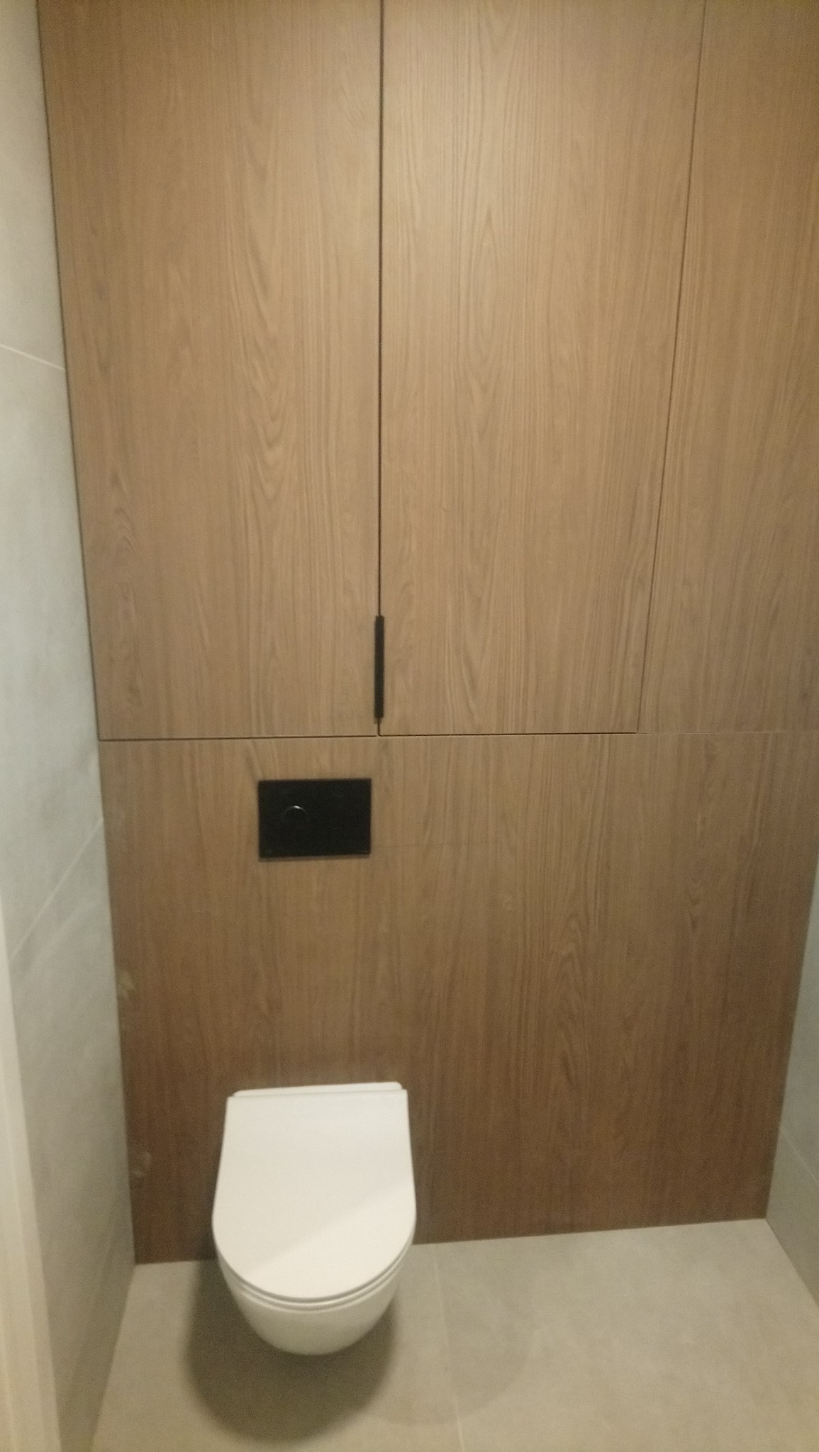 Nowoczesna toaleta podwieszana z drewnianą zabudową i czarnym panelem sterującym spłukiwaniem, widok z przodu, jasne płytki podłogowe i ścienne.