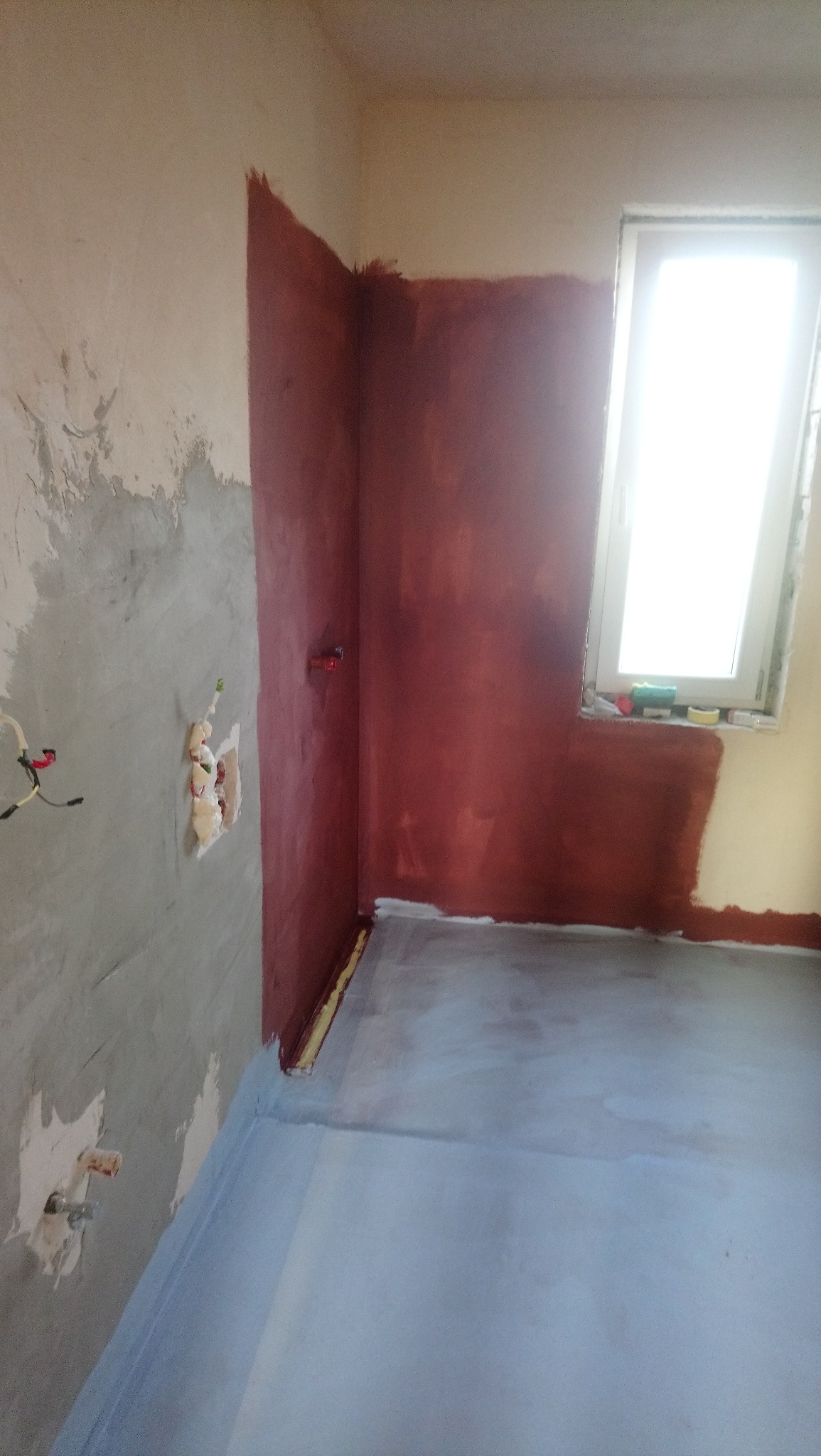 Przygotowanie ścian i podłogi w łazience: częściowo pomalowane ściany na bordowo, surowy beton na ścianie z instalacją, wylana podłoga, okno z widocznymi akcesoriami malarskimi.