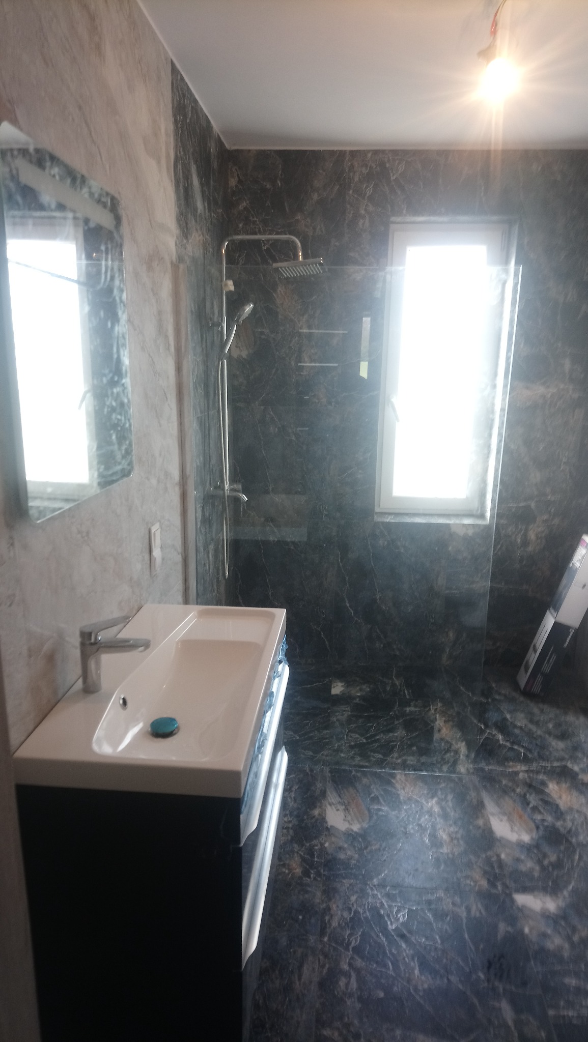 Wykończona łazienka z ciemnymi płytkami imitującymi marmur na ścianach i podłodze, kabina prysznicowa typu walk-in, umywalka nablatowa z szafką w kolorze granatowym, duże lustro na ścianie.