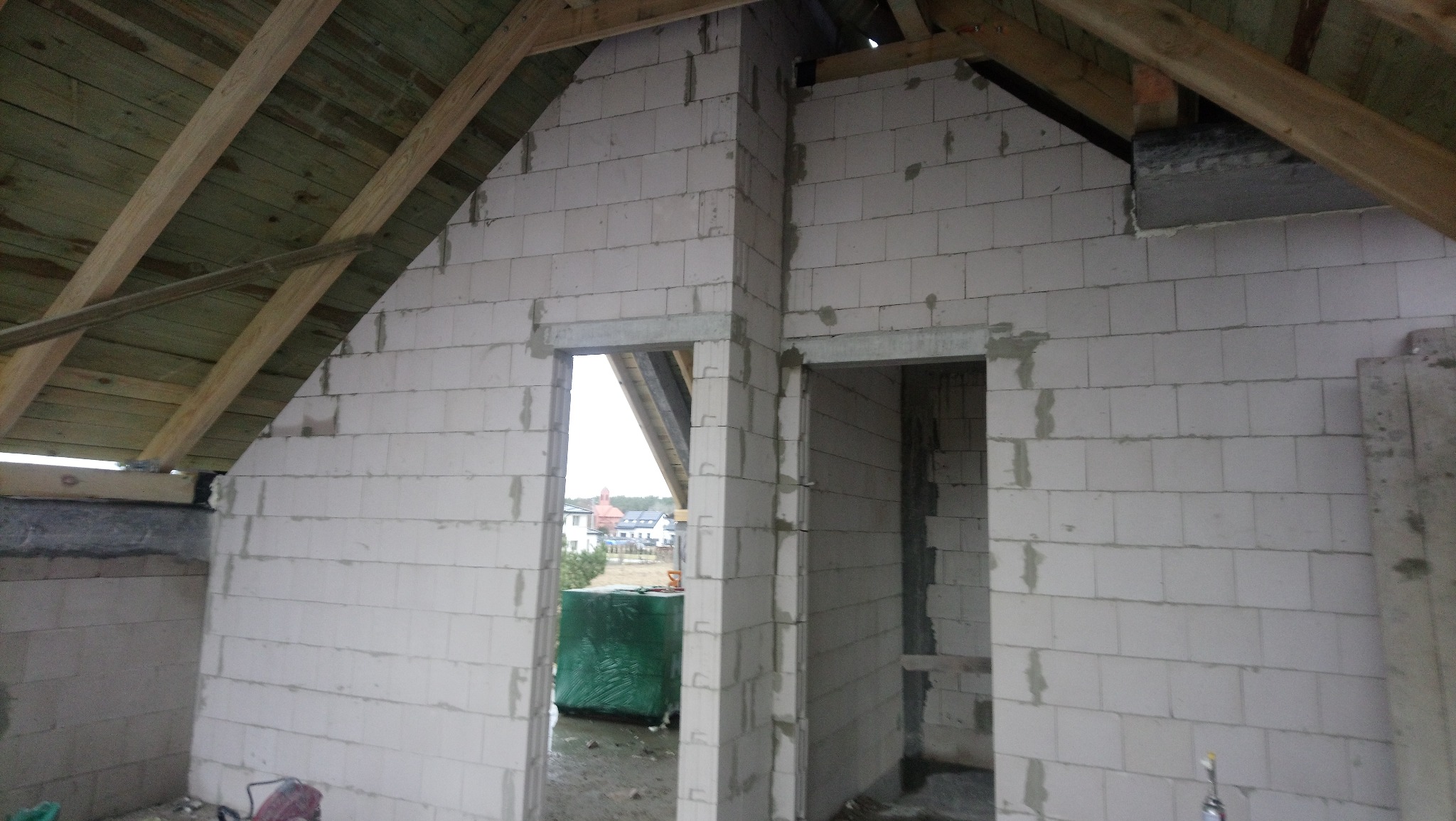 Surowy stan poddasza z widoczną konstrukcją dachu, ścianami z bloczków i przygotowanymi otworami na drzwi, z widokiem na zabudowę mieszkalną w tle.