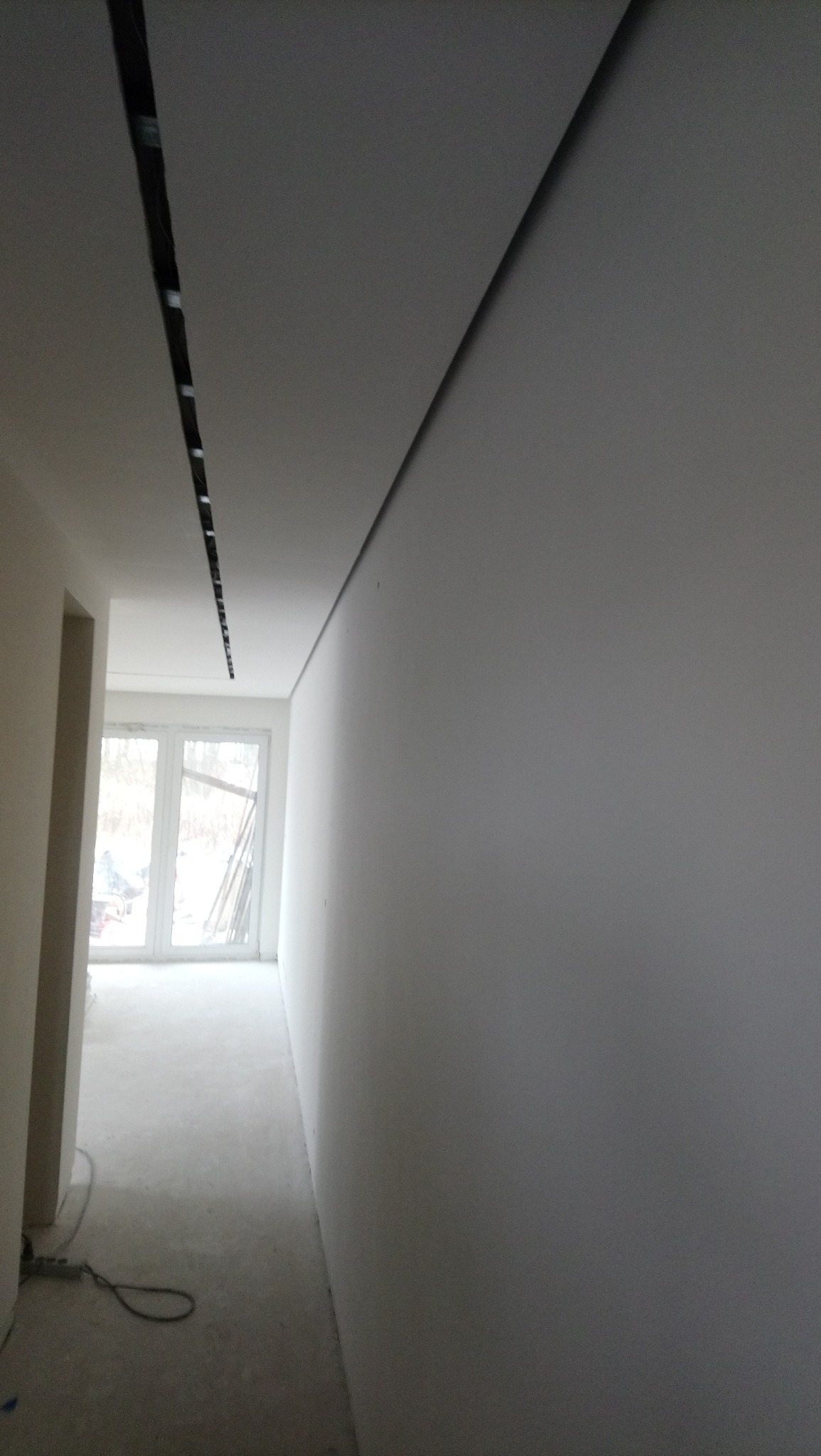 Korytarz w trakcie wykańczania, surowa ściana po gładzi, widoczne listwy oświetleniowe w suficie, okno z widokiem na zewnątrz.