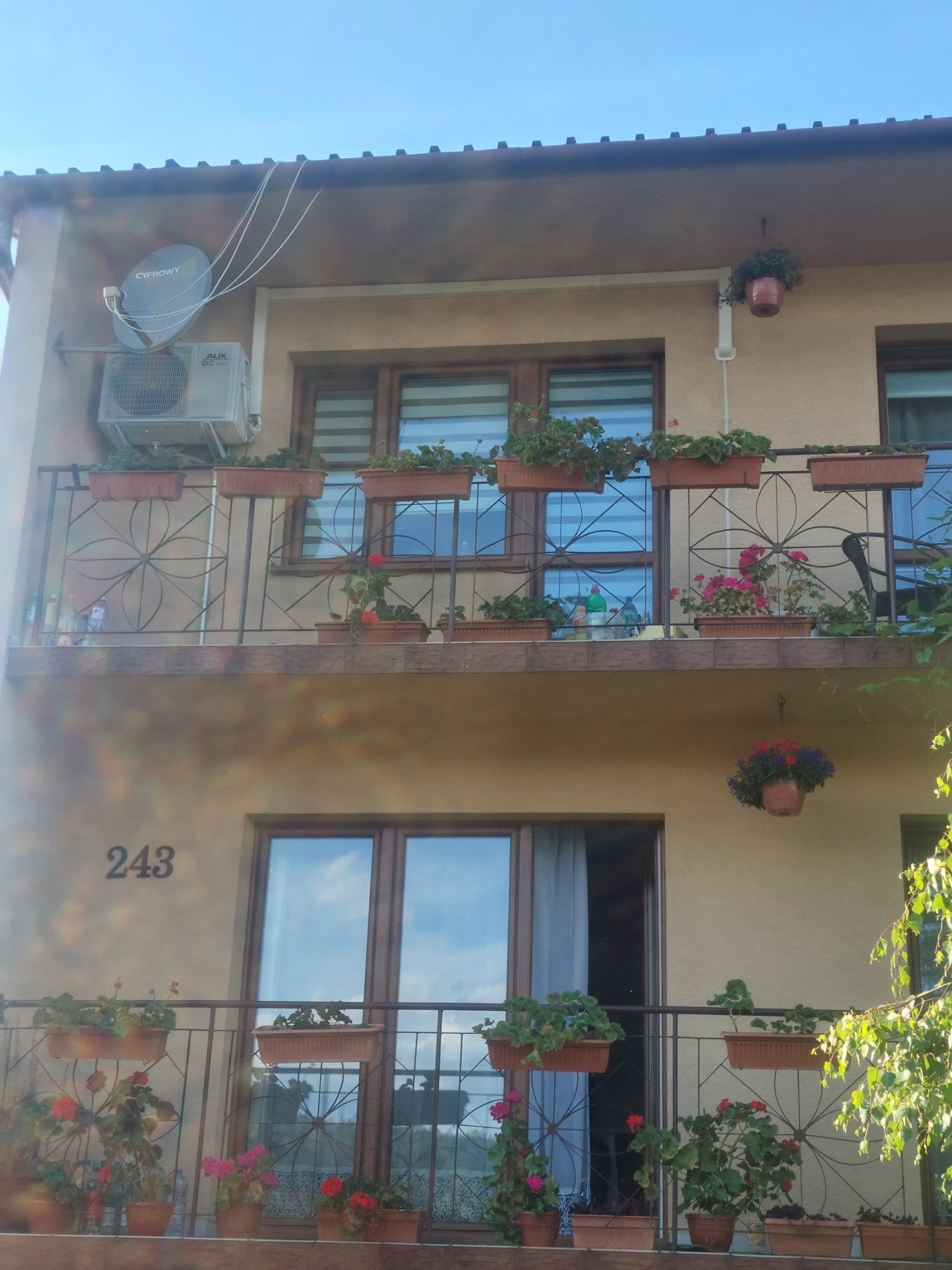 Elewacja dwupiętrowego domu z balkonami ozdobionymi kwiatami w donicach, klimatyzator na ścianie obok anteny satelitarnej