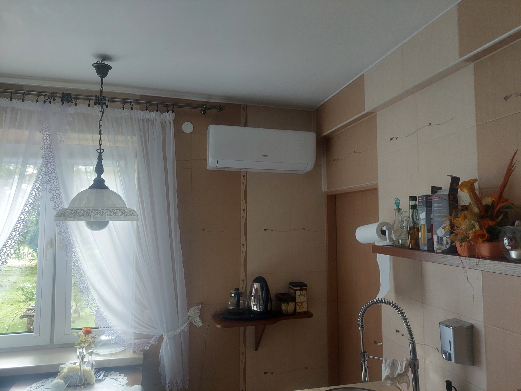 Biały klimatyzator AUX zamontowany na ścianie w kuchni z beżowymi panelami, widoczny fragment okna z firanką i lampą wiszącą.