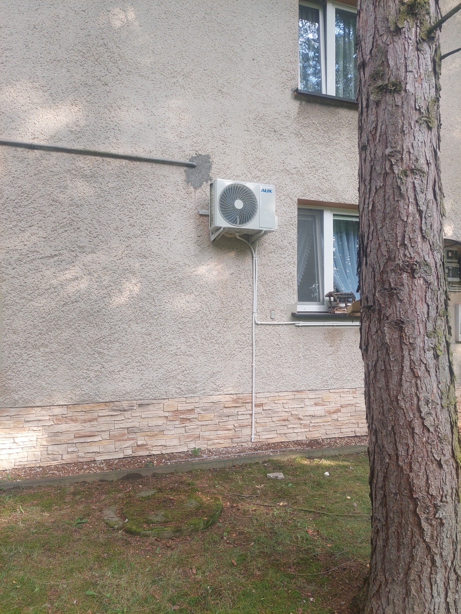 Zewnętrzna jednostka klimatyzatora AUX zamontowana na elewacji budynku z oknami, widoczna rura odprowadzająca skropliny, obok pień drzewa.