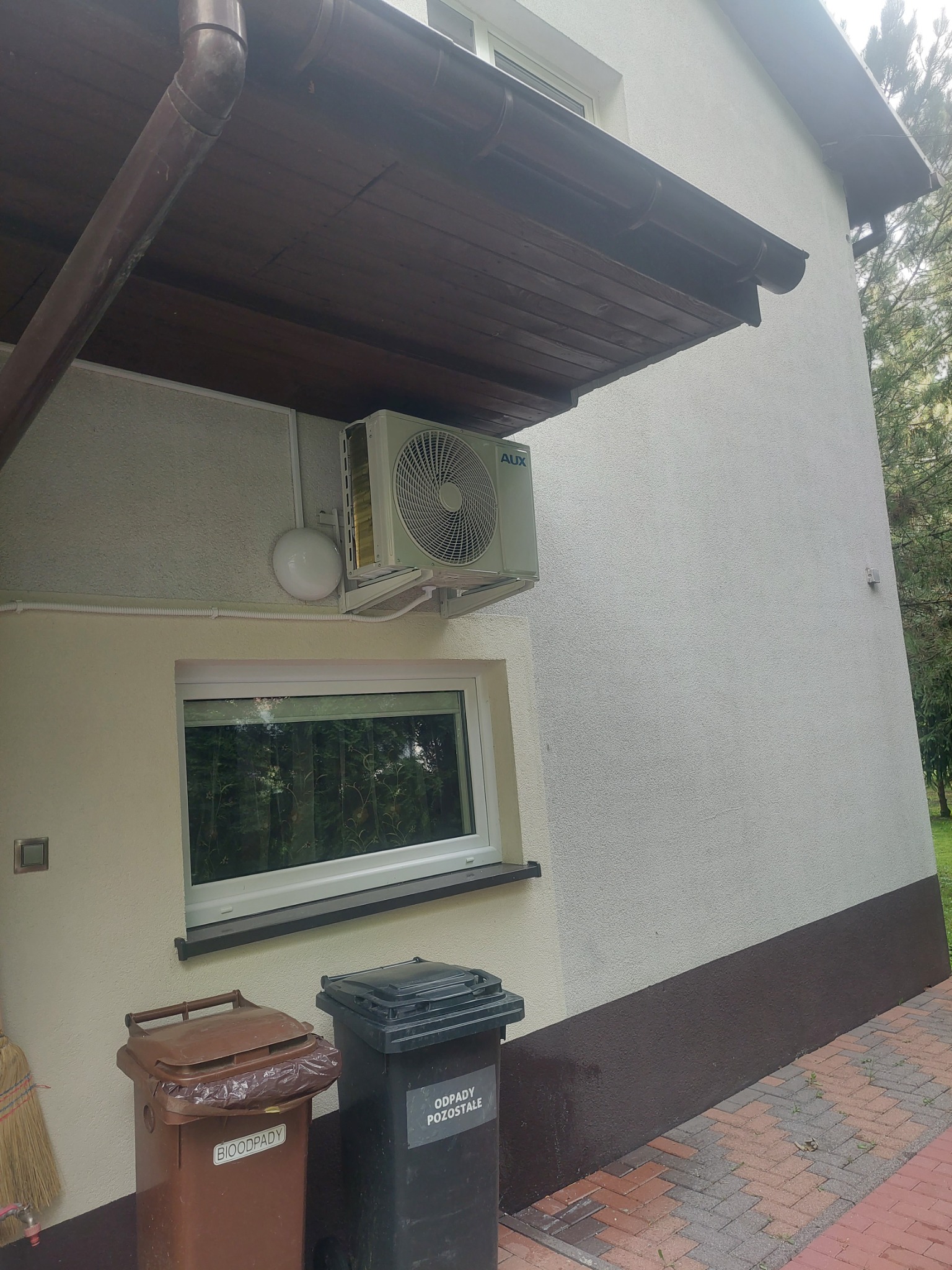 Zewnętrzna jednostka klimatyzacji AUX zamontowana na ścianie domu, widoczna instalacja elektryczna i odpływ skroplin, obok okna i koszy na śmieci.