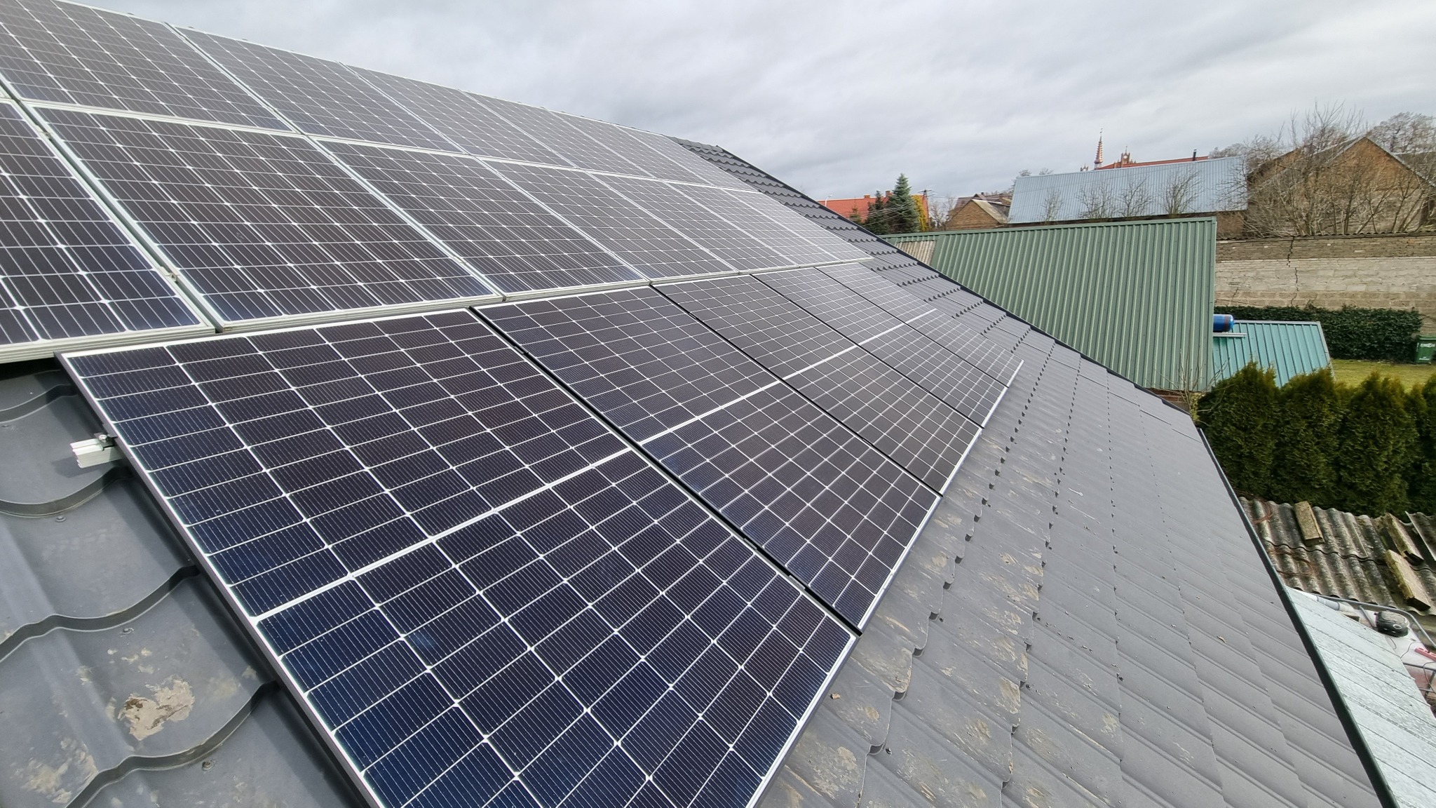 Panele słoneczne zamontowane na dachu z szarej dachówki, widok z góry, w tle zabudowania gospodarcze i kościół