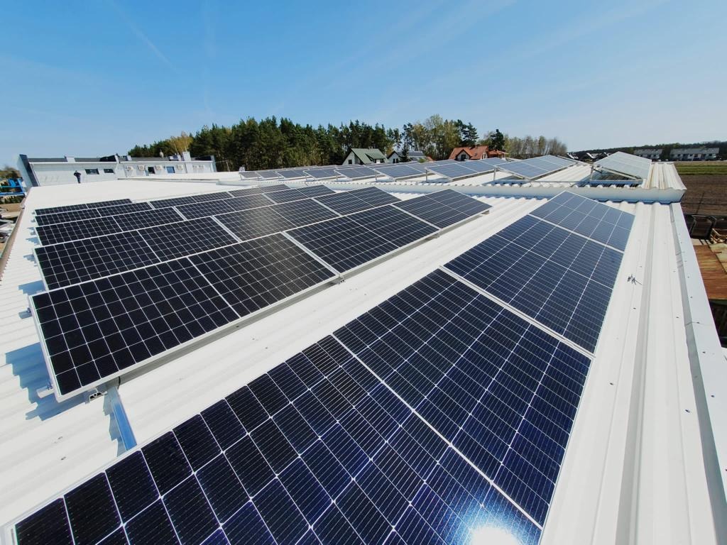 Rząd paneli słonecznych na białym, metalowym dachu budynku w słoneczny dzień, widoczne drzewa w tle.