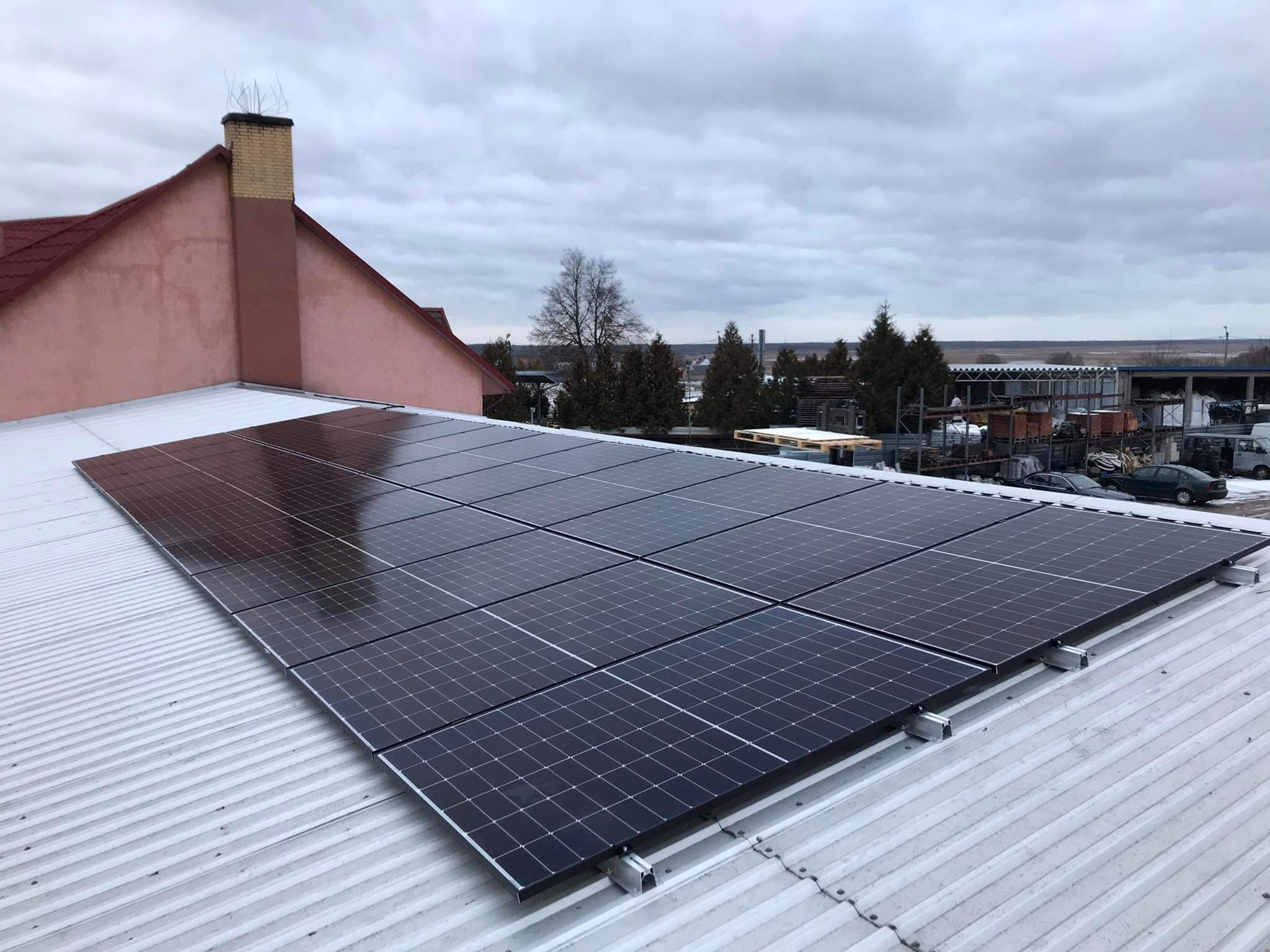 Panele słoneczne zamontowane na blaszanym dachu budynku z kominem, widok z góry w pochmurny dzień.