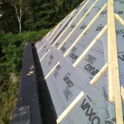 Konstrukcja dachu w trakcie budowy, widoczne drewniane łaty i membrana dachowa z nadrukiem VAXO 3XL, w tle zieleń drzew.