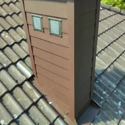 Nowoczesna obudowa komina wentylacyjnego w kolorze brązowym, z dwoma kratkami wentylacyjnymi, dopasowana do pokrycia dachowego z dach&oacute;wki ceramicznej.