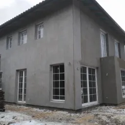 Surowy, dwukondygnacyjny dom murowany w trakcie wykonywania elewacji zewnętrznej, z widocznymi rusztowaniami i stosem desek na ośnieżonym terenie.