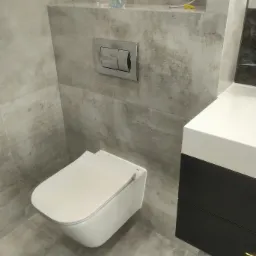 Nowoczesna toaleta podwieszana z szarymi płytkami imitującymi kamień, chromowanym przyciskiem spłukującym i umywalką z szafką w kolorze czarnym.