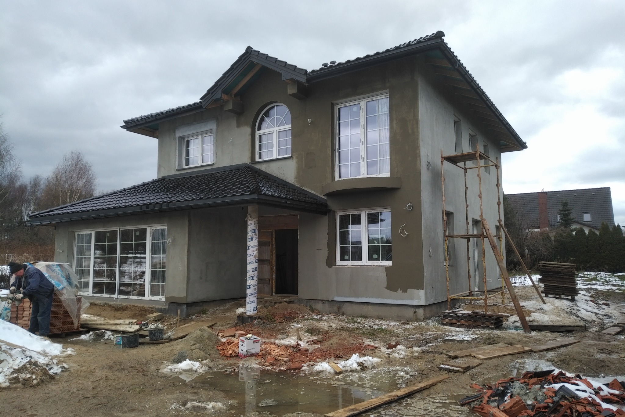 Dwukondygnacyjny dom murowany w trakcie budowy, z czarnym dachem, częściowo otynkowany, z widocznym rusztowaniem i pracownikiem układającym cegły na zewnątrz.