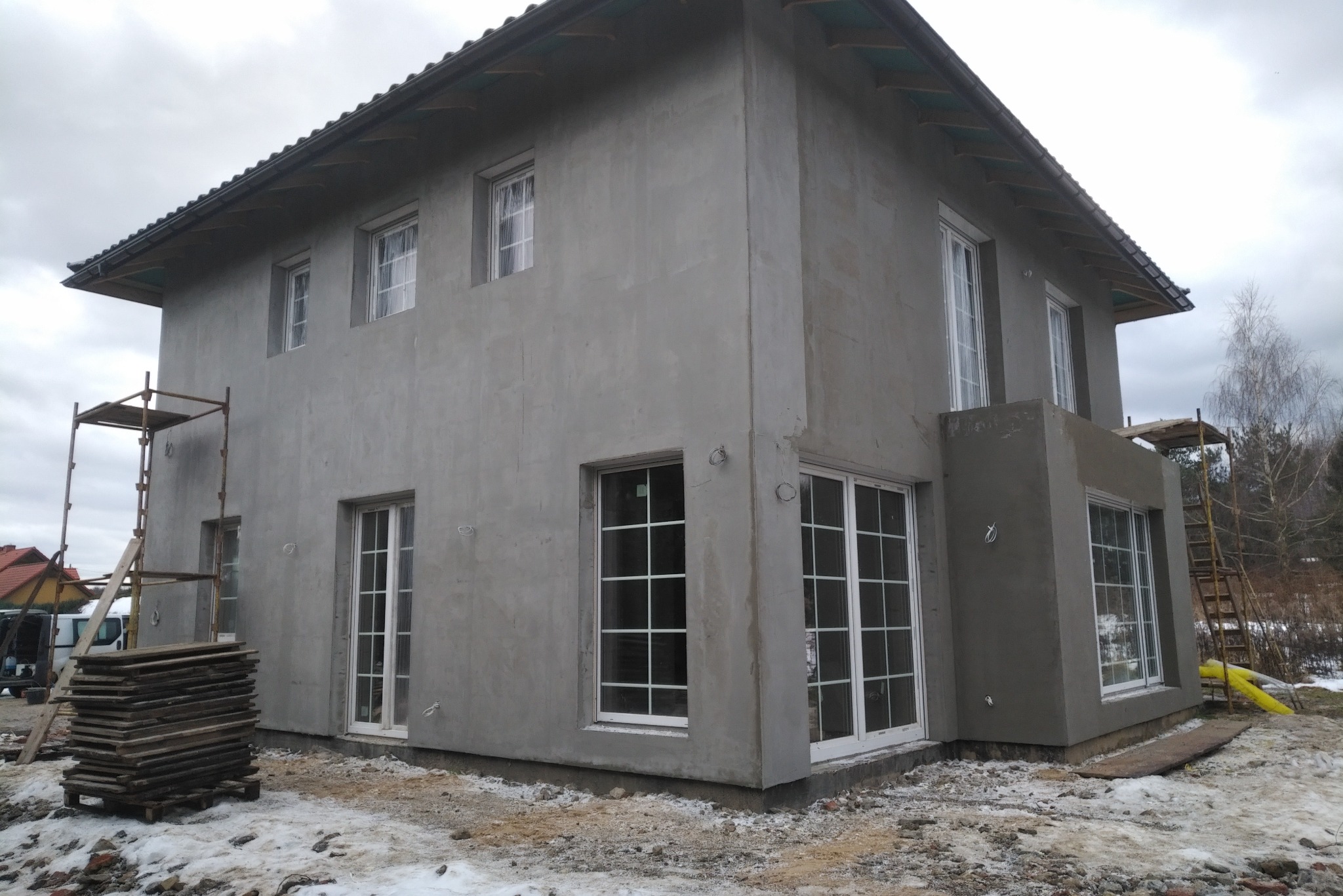 Surowy, dwukondygnacyjny dom murowany w trakcie wykonywania elewacji zewnętrznej, z widocznymi rusztowaniami i stosem desek na ośnieżonym terenie.