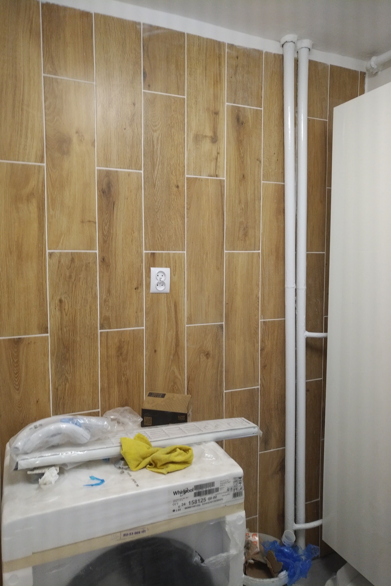 Ściana wyłożona brązowymi płytkami imitującymi drewno, z widocznym gniazdkiem elektrycznym, białymi rurami i fragmentem białej szafki, z pralką i żółtą ścierką na pierwszym planie.