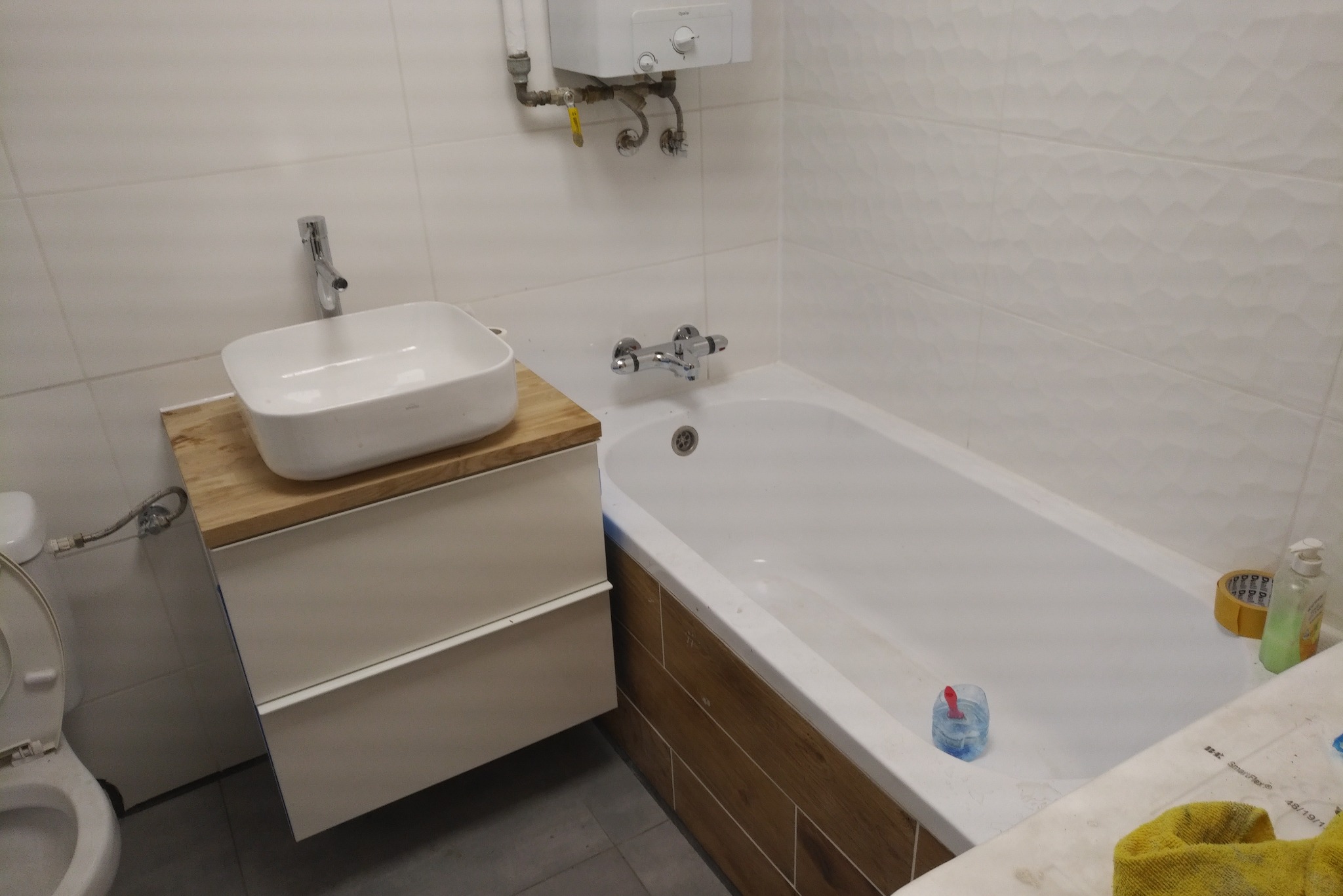 Nowoczesna łazienka z białą wanną obudowaną drewnopodobnymi płytkami, umywalką nablatową na szafce z drewnianym blatem, oraz białymi płytkami ściennymi z delikatną fakturą.