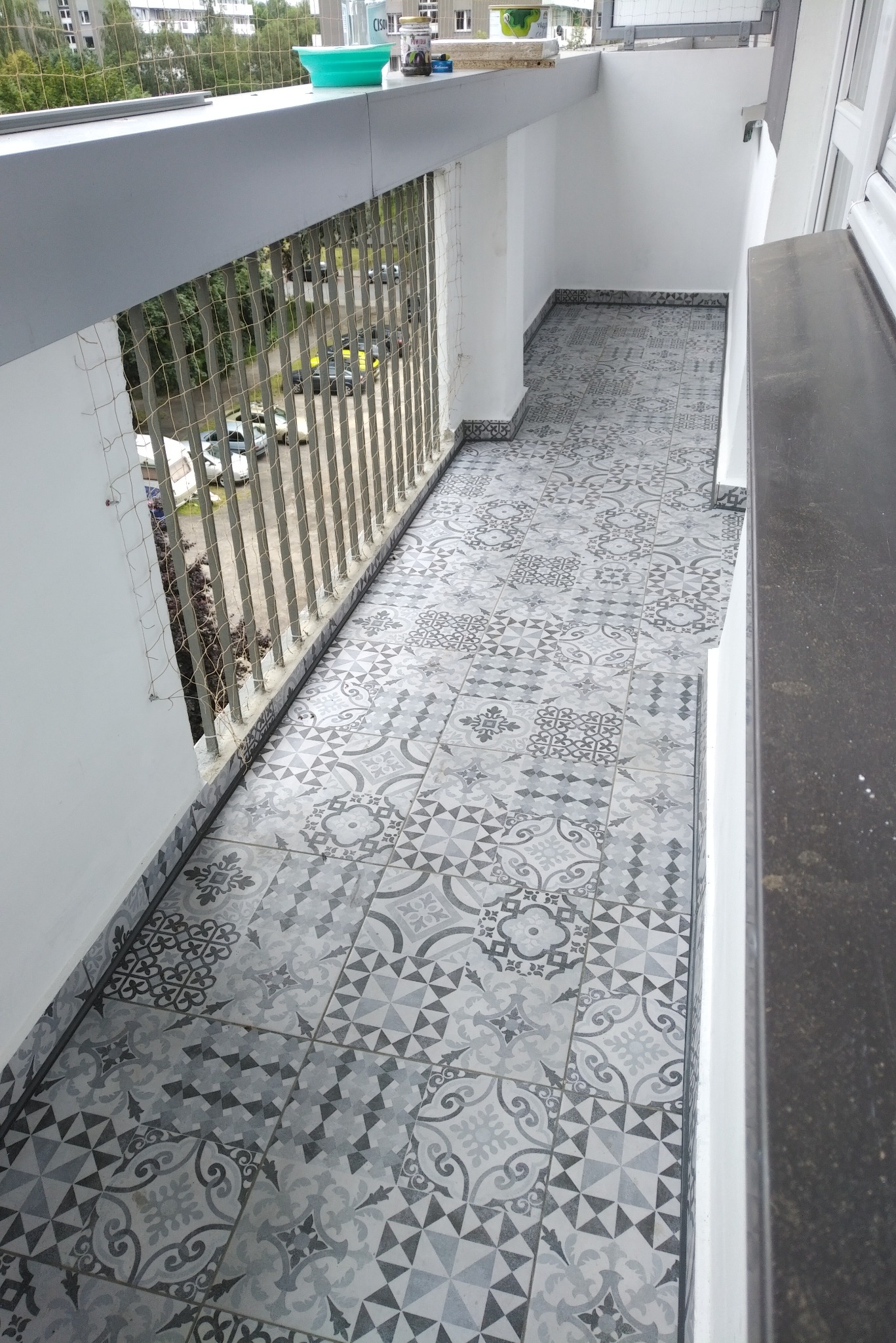 Wykończony balkon z szarą, wzorzystą podłogą z płytek ceramicznych, zabezpieczony siatką ochronną, widok z góry na fragment osiedla.