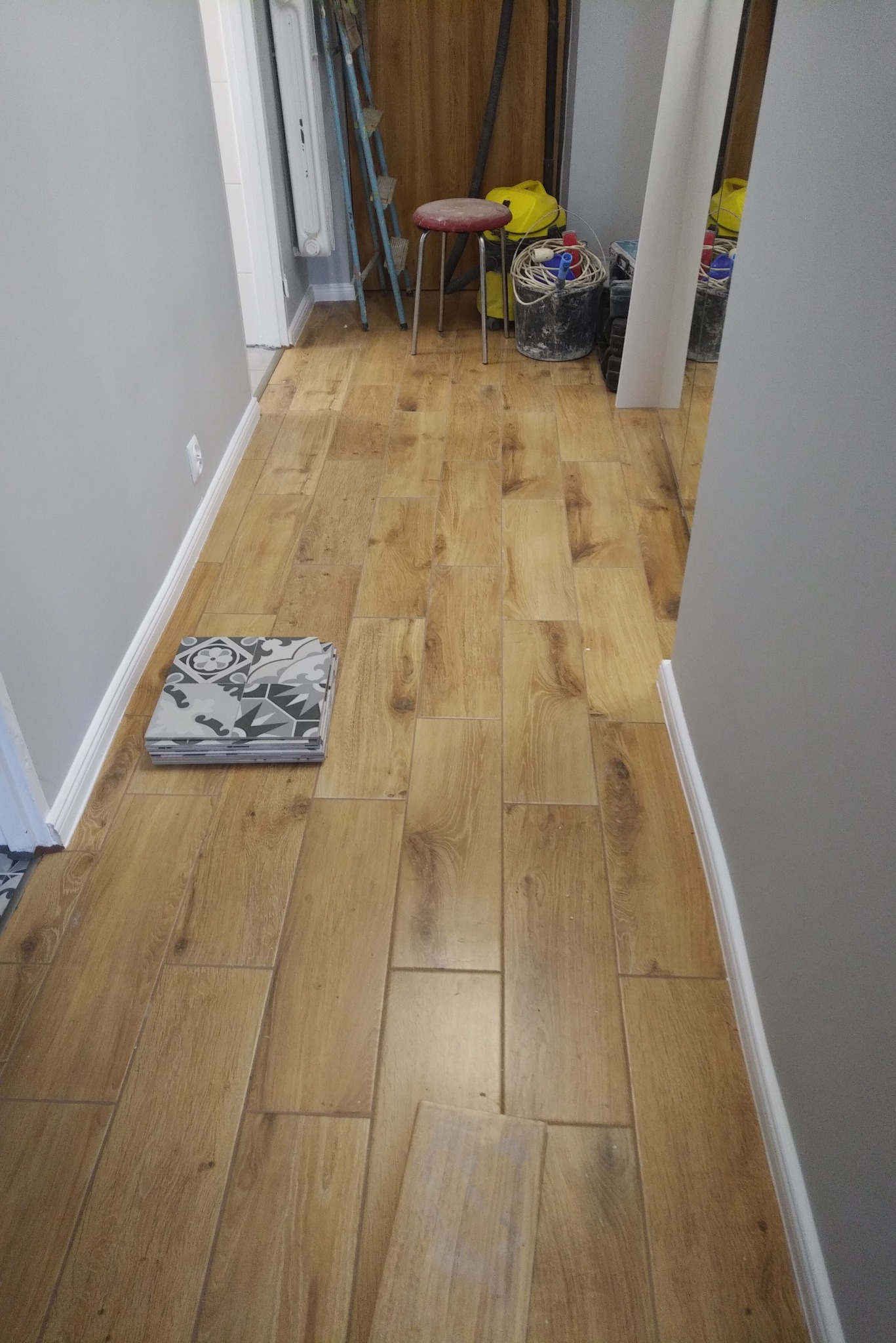 Ułożona podłoga z paneli drewnopodobnych w korytarzu, obok ściany leży stos płytek dekoracyjnych, w tle widoczne narzędzia i drabina.