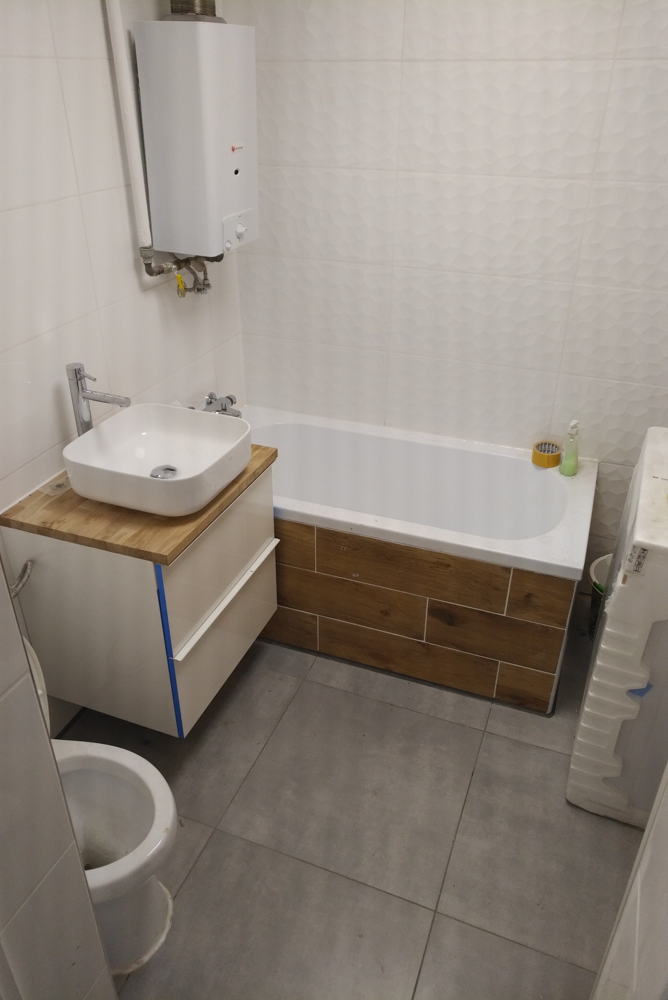 Nowoczesna łazienka z białą ceramiką, drewnianym blatem pod umywalką i obudową wanny imitującą drewno, grzejnik gazowy na ścianie z dekoracyjnymi płytkami.