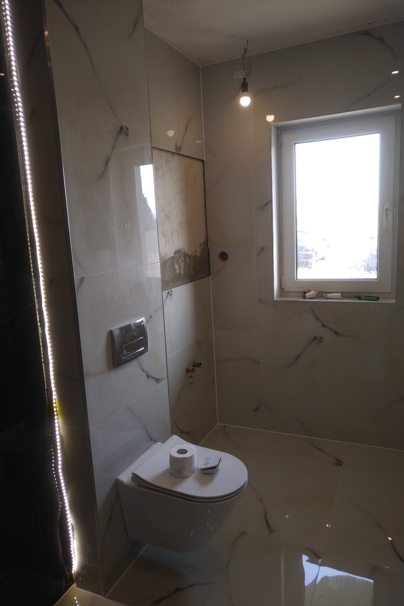 Wnętrze łazienki w trakcie remontu, widoczne płytki imitujące marmur, wisząca miska WC z deską, fragment ściany bez płytek, okno i oświetlenie podsufitowe.