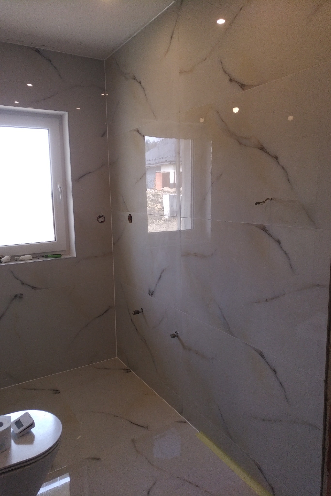 Wykończona łazienka z płytkami imitującymi marmur na ścianach i podłodze, widoczne przygotowane punkty pod armaturę, okno z widokiem na budynek w tle.