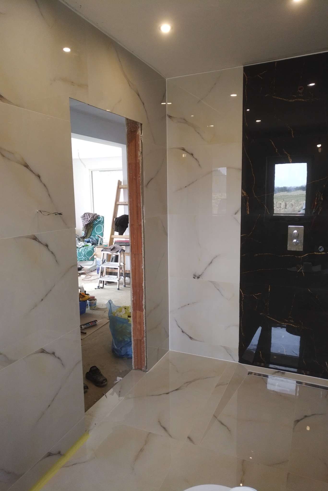 Wykończenie łazienki w trakcie: białe i czarne płytki imitujące marmur na ścianach i podłodze, widoczny otwór drzwiowy z surowym wykończeniem, drabina malarska i narzędzia w tle.