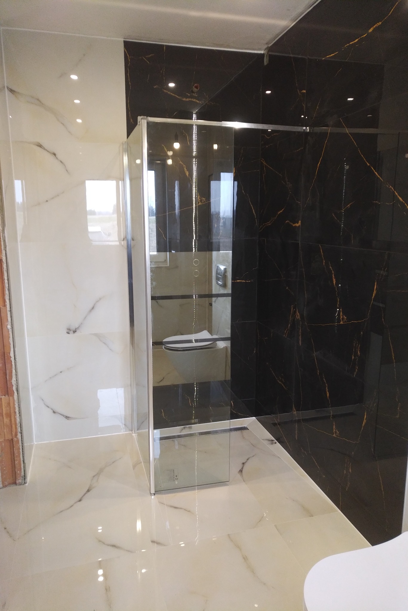 Nowoczesna kabina prysznicowa ze szklanymi ściankami na tle czarno-złotych i biało-marmurkowych płytek w wykończonej łazience, widoczne oświetlenie punktowe.