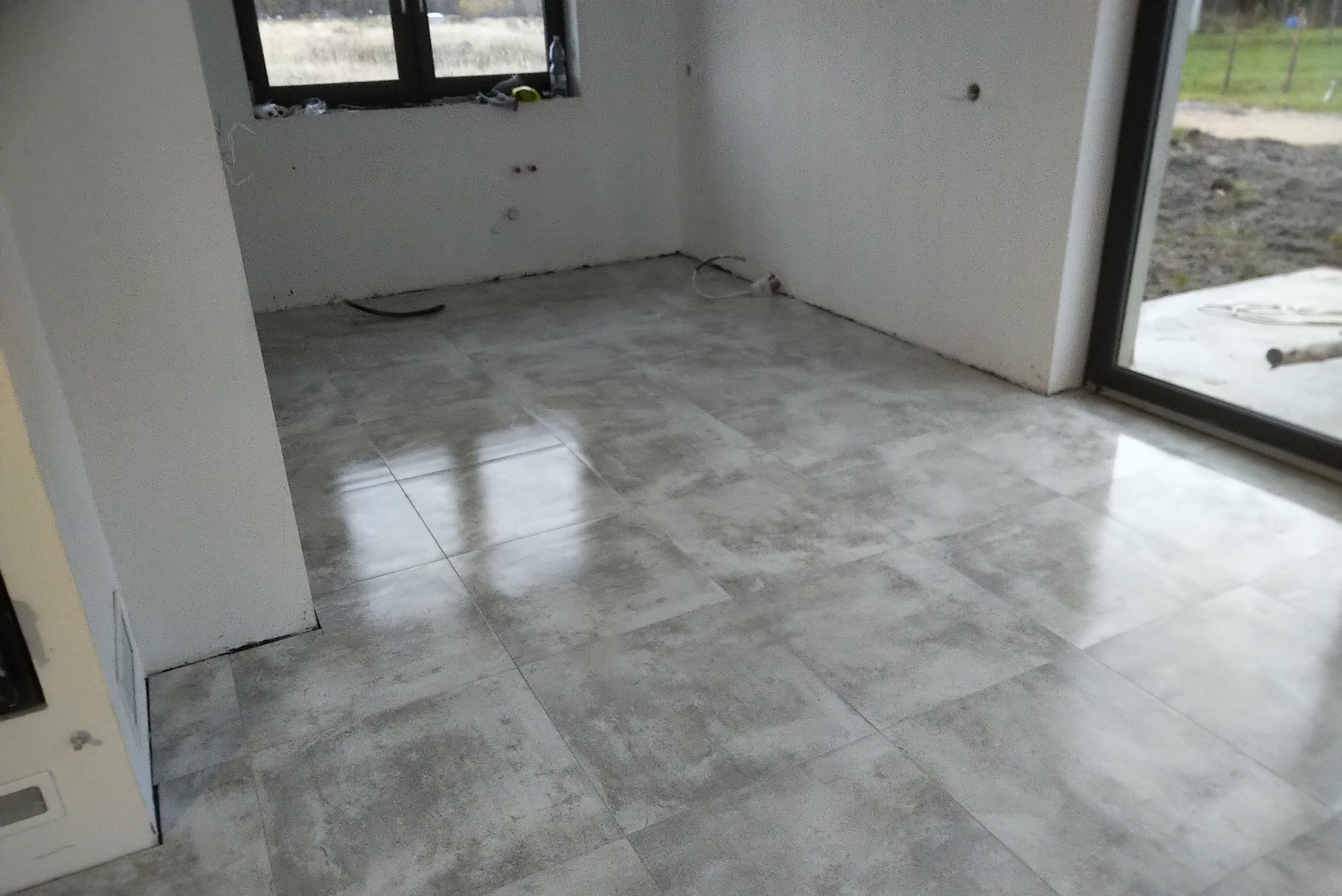 Wnętrze w trakcie wykańczania: szare płytki podłogowe imitujące beton, surowe ściany z widocznymi instalacjami elektrycznymi, okno z widokiem na zewnątrz.