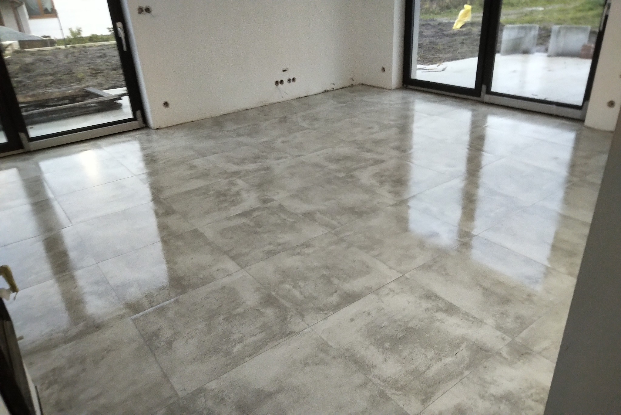 Wykończone wnętrze domu z błyszczącą, szarą podłogą z płytek ceramicznych, duże okna przesuwne z widokiem na ogród i przygotowane punkty elektryczne na ścianie.