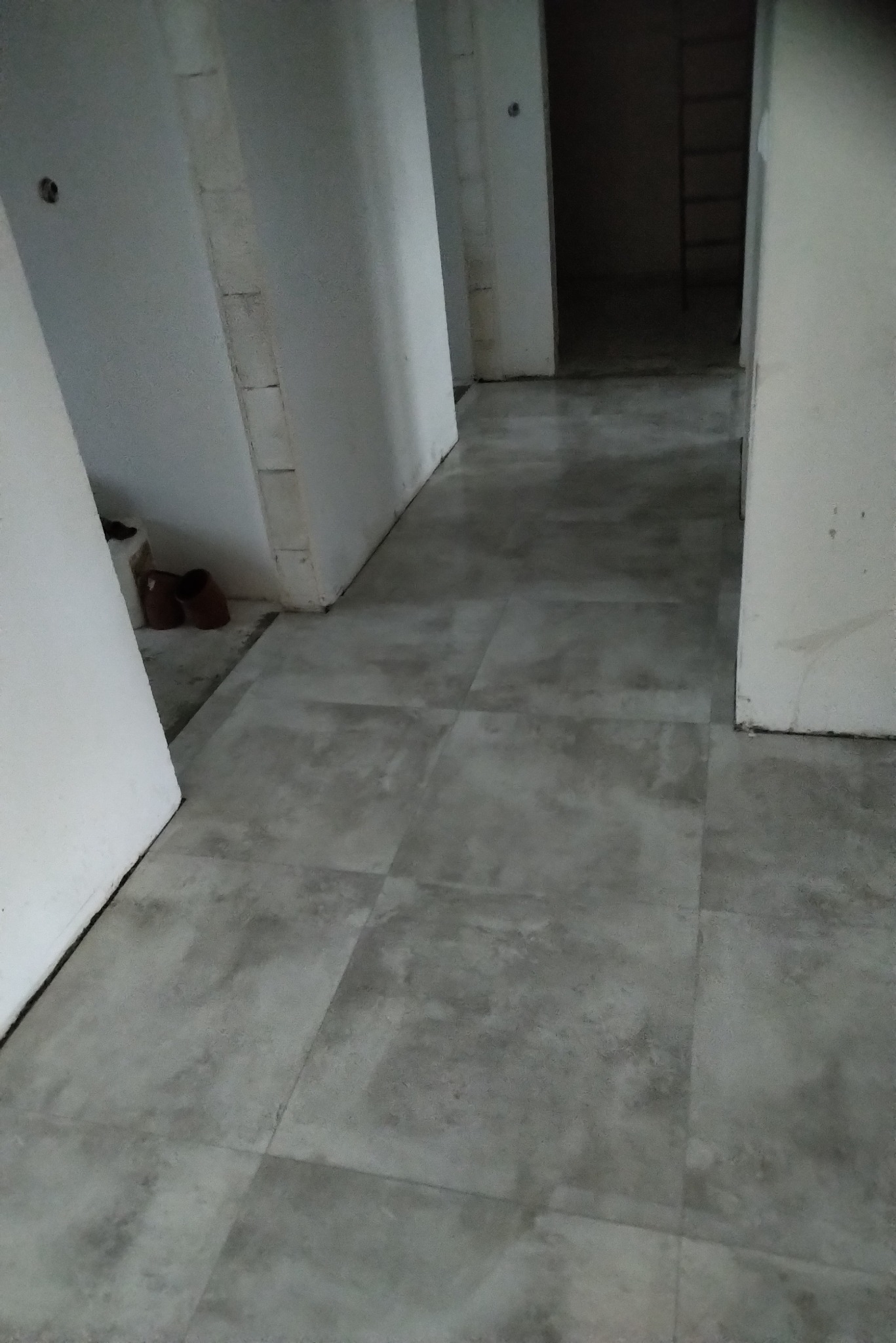 Wnętrze w trakcie budowy: szare płytki podłogowe ułożone w kwadraty, widoczne białe ściany z surowego muru i drabina w głębi, para brązowych butów przy ścianie.