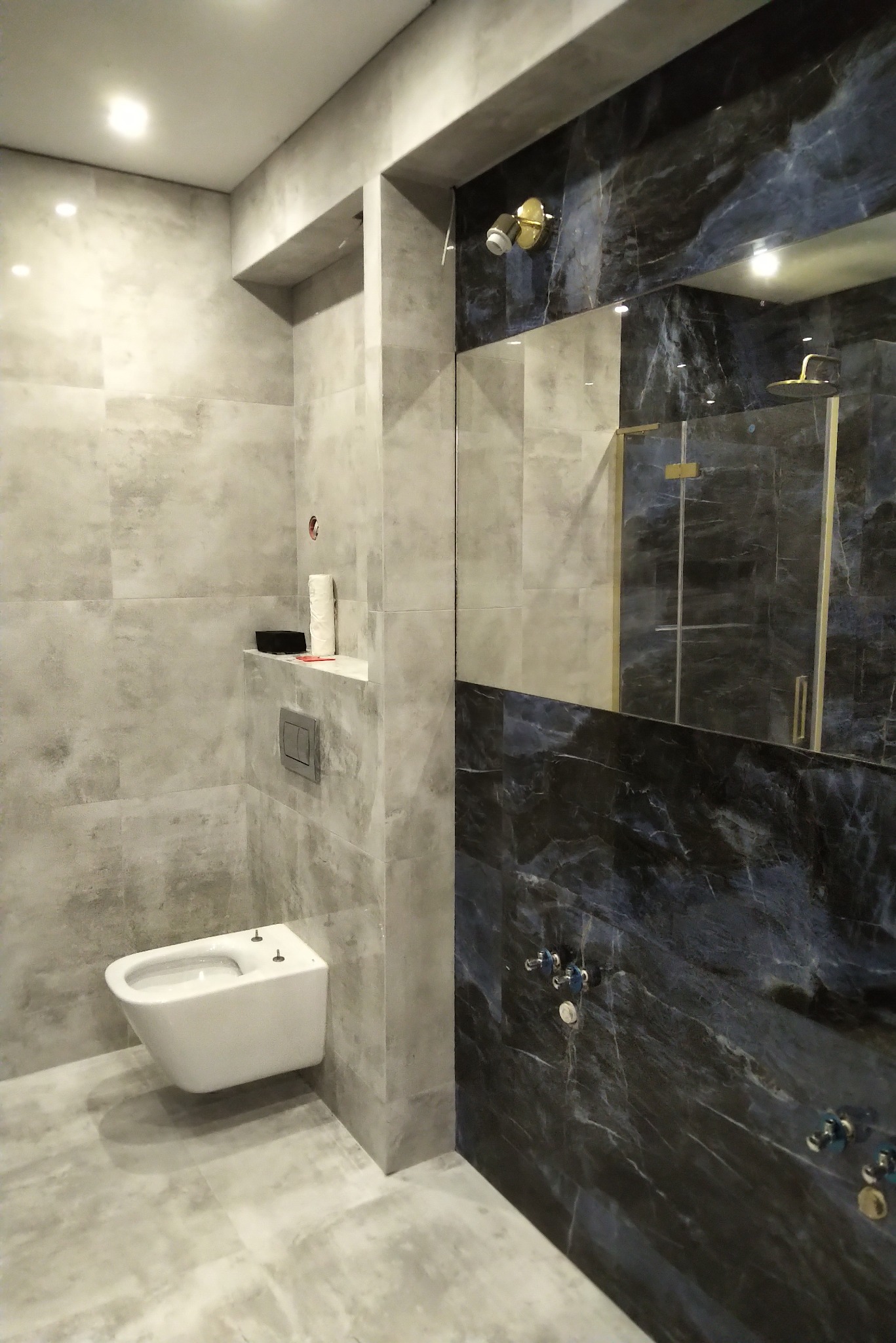 Nowoczesna łazienka z marmurową ścianą, podwieszaną toaletą i kabiną prysznicową ze złotymi detalami, oświetlona punktowo.