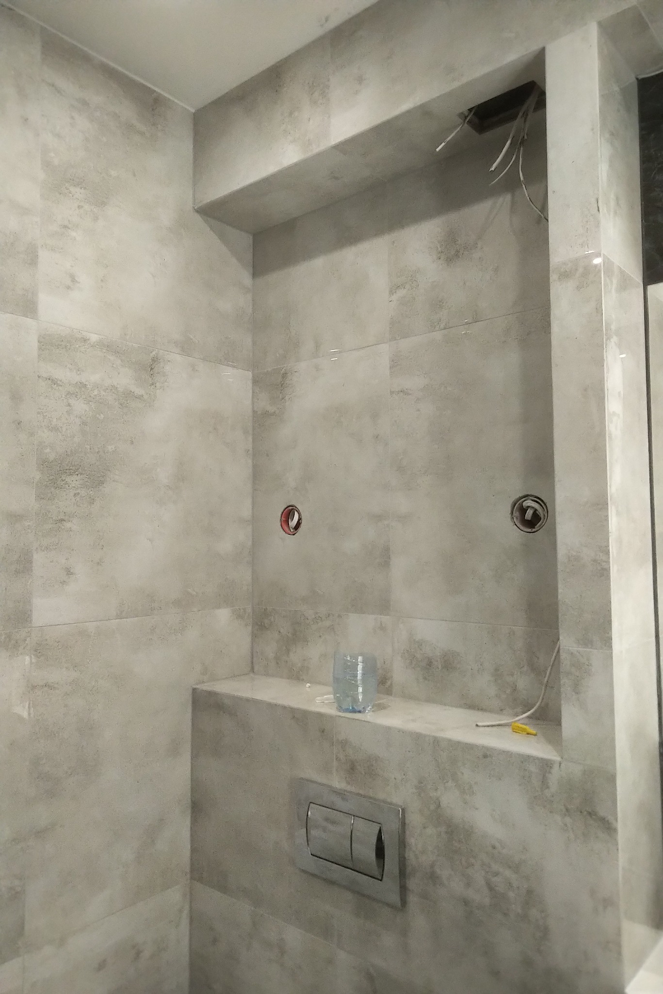 Wnętrze łazienki w trakcie wykańczania z szarą, wzorzystą glazurą, widoczne otwory na baterie, wystające przewody elektryczne, spłuczka podtynkowa oraz plastikowa butelka na półce.