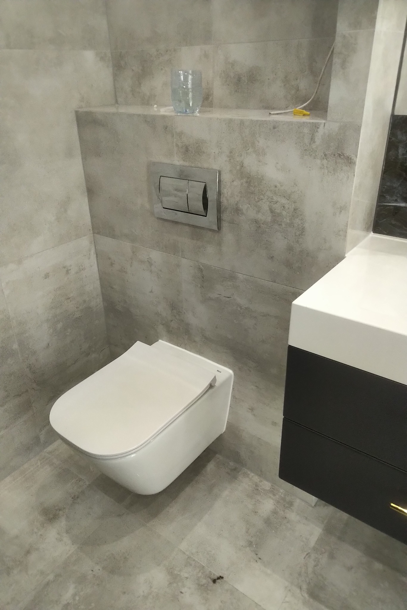 Nowoczesna toaleta podwieszana z szarymi płytkami imitującymi kamień, chromowanym przyciskiem spłukującym i umywalką z szafką w kolorze czarnym.