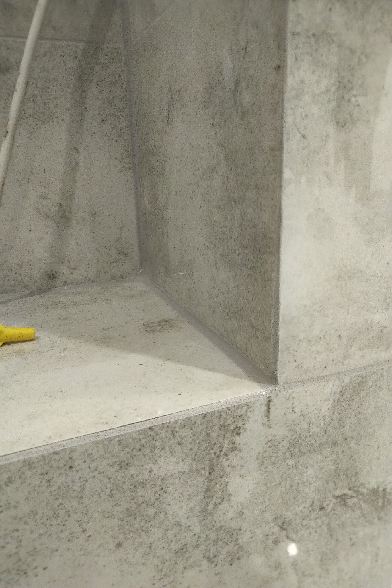 Wnęka w ścianie wyłożona szarą, wzorzystą glazurą, z widocznym żółtym elementem narzędzia po lewej stronie.