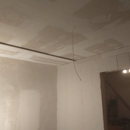 PawMat - Wnętrze w trakcie prac wykończeniowych: ściany i sufit częściowo pokryte gładzią, widoczne listwy montażowe i przewód elektryczny, w tle widoczne schody i drabina.