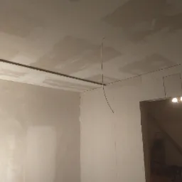 Wnętrze w trakcie prac wykończeniowych: ściany i sufit częściowo pokryte gładzią, widoczne listwy montażowe i przewód elektryczny, w tle widoczne schody i drabina.