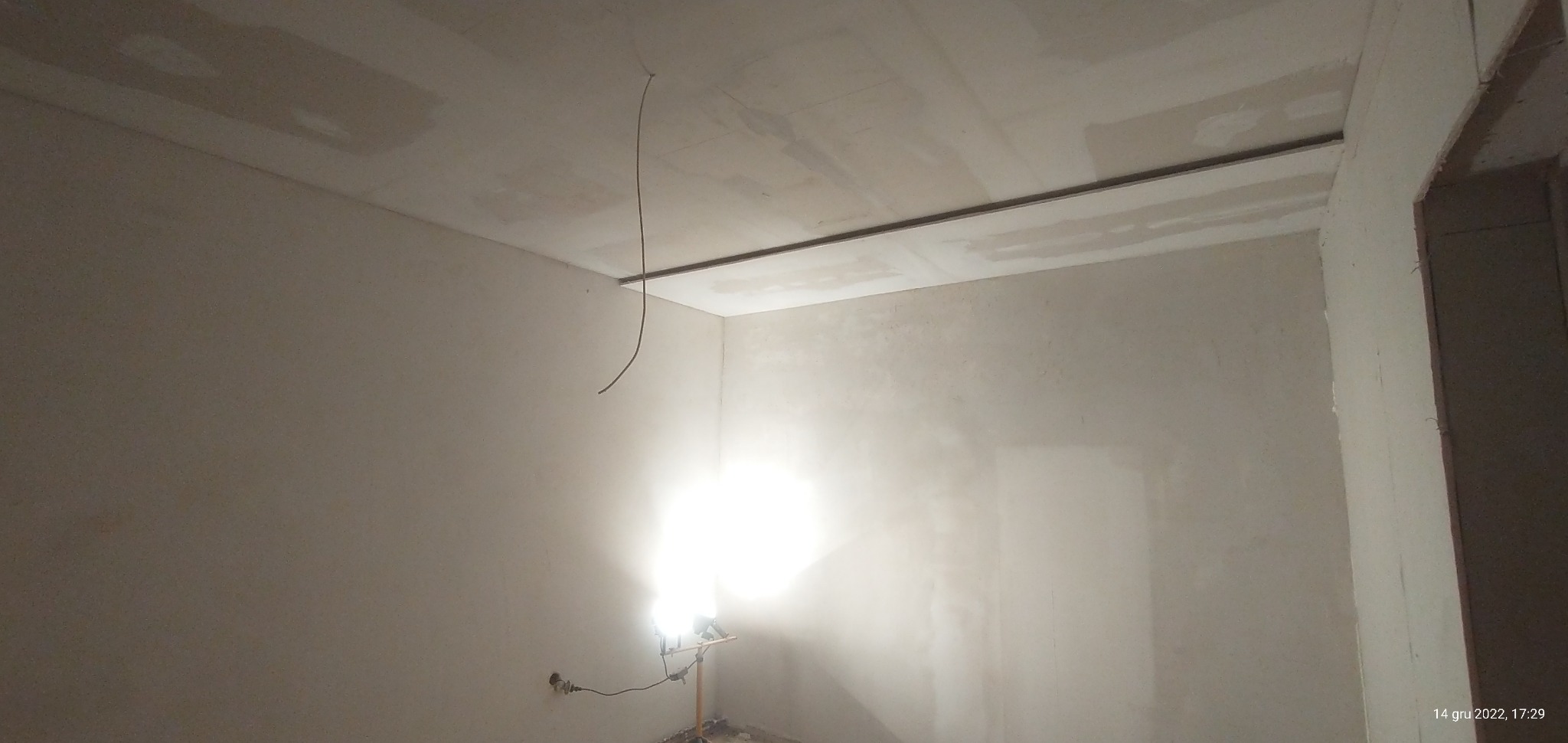 Wnętrze w trakcie prac wykończeniowych: ściany po tynkowaniu, widoczna instalacja elektryczna, sufit z zamontowanymi płytami gipsowo-kartonowymi, oświetlone lampą budowlaną.