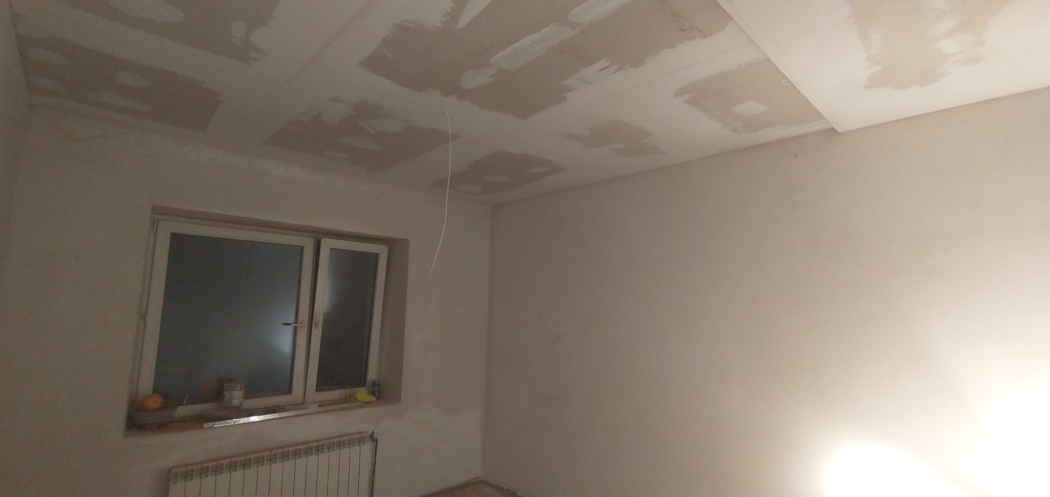Pokój w trakcie prac wykończeniowych: ściany i sufit częściowo pokryte gładzią, widoczne okno z parapetem, grzejnik pod oknem i wystający kabel ze stropu.