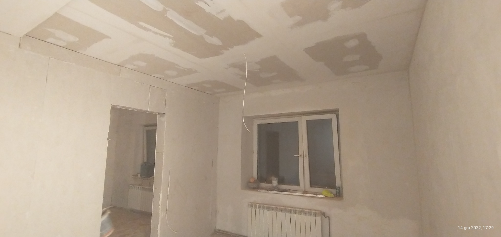 Wnętrze pokoju w trakcie remontu: widoczne ściany pokryte gładzią w trakcie nakładania, sufit z widocznymi fragmentami gładzi, niezakryty kabel zwisający z sufitu, okno z grzejnikiem pod spodem...