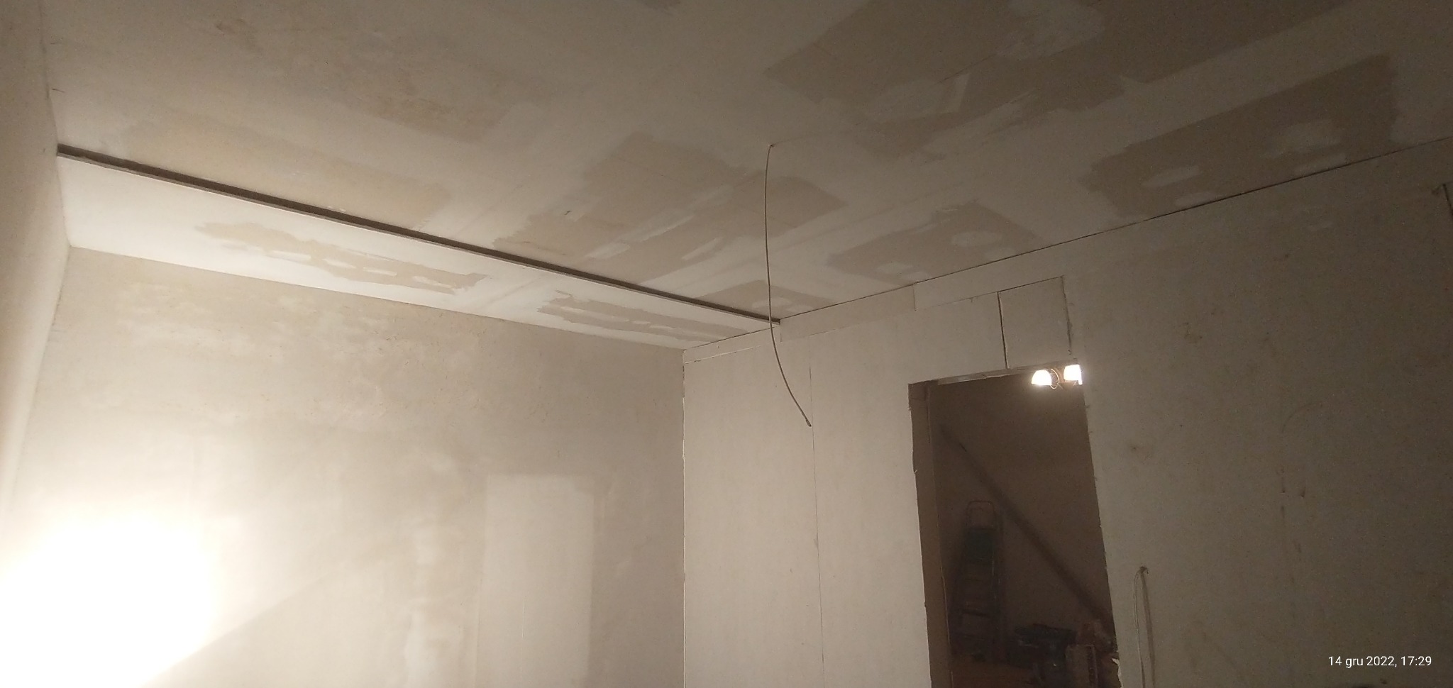 Wnętrze w trakcie prac wykończeniowych: ściany i sufit częściowo pokryte gładzią, widoczne listwy montażowe i przewód elektryczny, w tle widoczne schody i drabina.