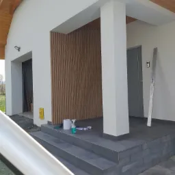 Nowoczesne wejście do domu z drewnianą okładziną ścienną, widoczne narzędzia i akcesoria przygotowane do montażu, poziomica oparta o ścianę, szary kolor płytek na schodach i podłodze.