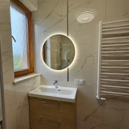 Wykończona łazienka z okrągłym lustrem z podświetleniem LED, umywalką nablatową z szafką z drewnopodobnym fornirem, grzejnikiem drabinkowym i marmurowymi płytkami ściennymi.