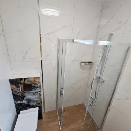 Nowoczesna łazienka z kabiną prysznicową ze srebrnymi profilami, podwieszaną toaletą z czarną obudową imitującą kamień oraz płytkami imitującymi biały marmur na ścianach i drewnopodobną podłogą.