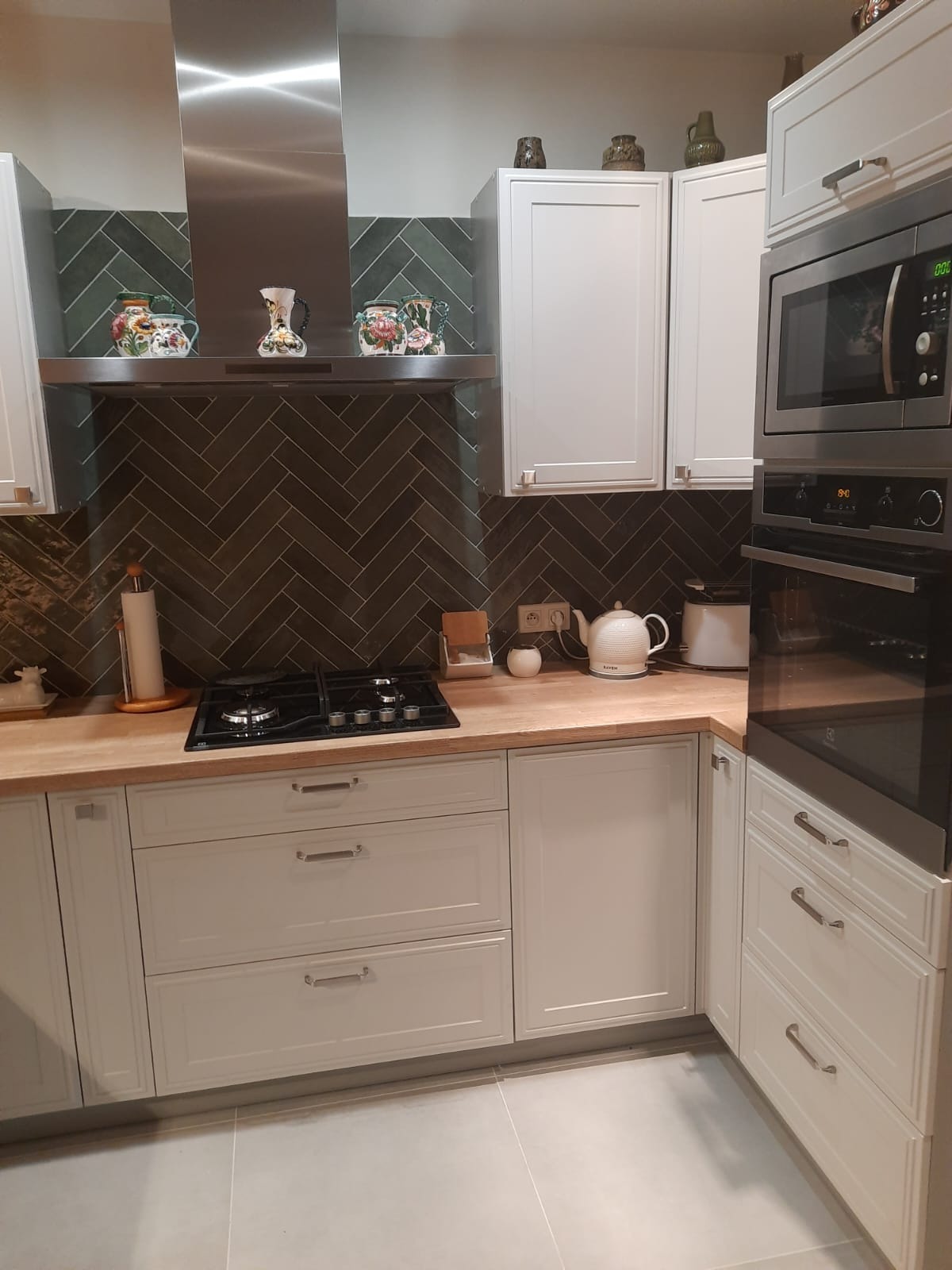 Nowoczesna kuchnia z białymi szafkami, drewnianym blatem i ciemną, ułożoną w jodełkę glazurą. Na blacie kuchenka gazowa, czajnik i drobne dekoracje. Nad kuchenką stalowy okap, a nad nim ozdobne...