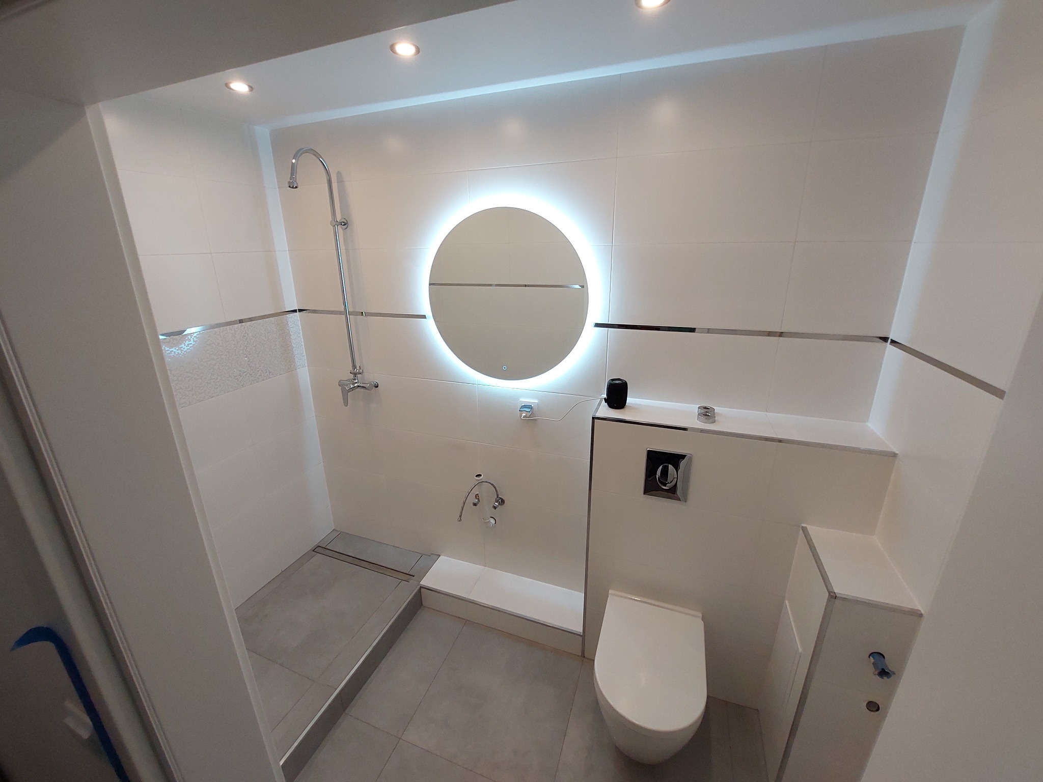 Nowoczesna łazienka z białymi płytkami, okrągłym lustrem z podświetleniem LED, kabiną prysznicową typu walk-in z odpływem liniowym i podwieszaną toaletą z zabudową.