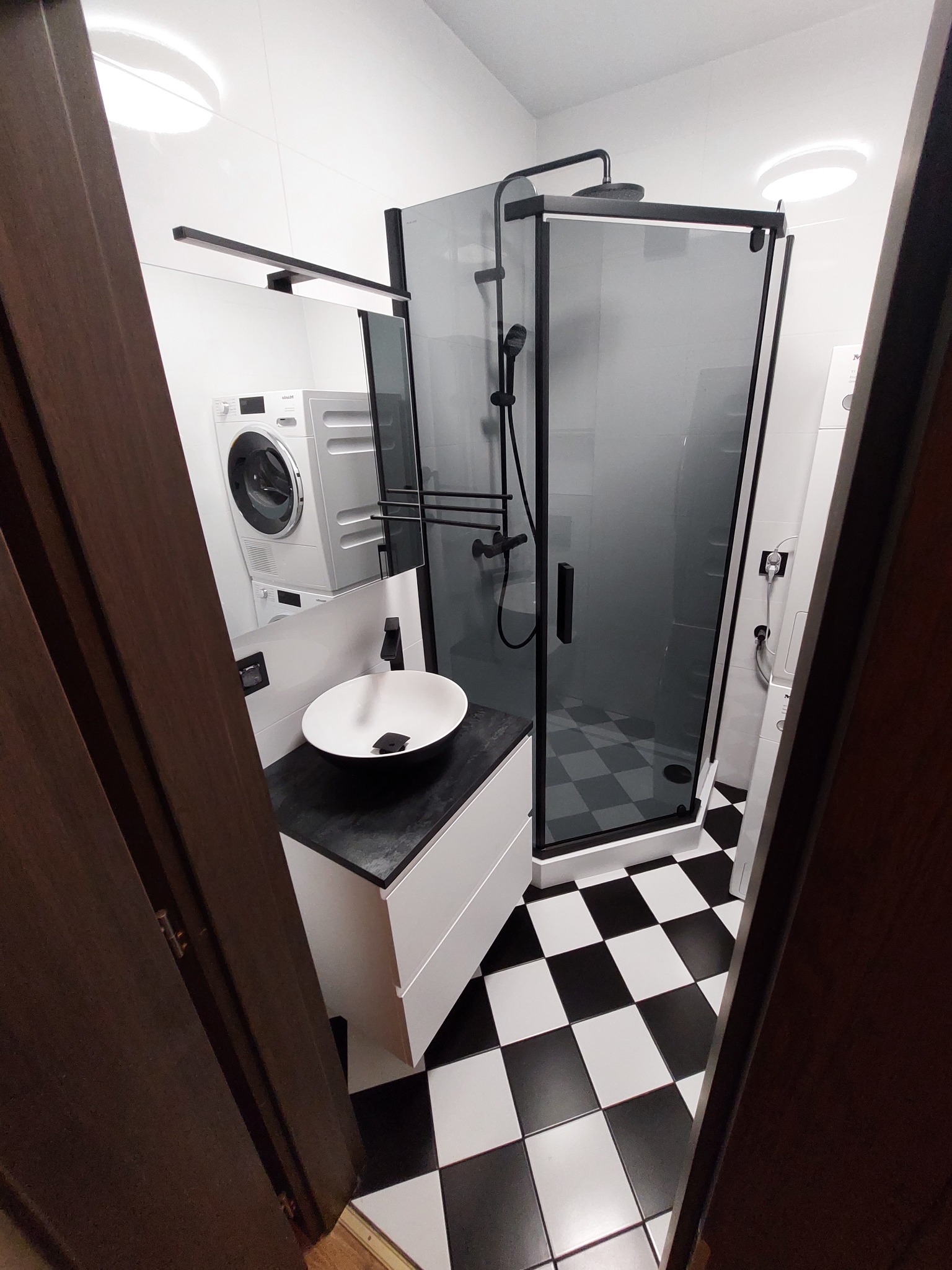 Nowoczesna łazienka z kabiną prysznicową typu walk-in z czarnymi profilami, umywalką nablatową na białej szafce z czarnym blatem, pralką i suszarką w zabudowie oraz biało-czarną szachownicą...