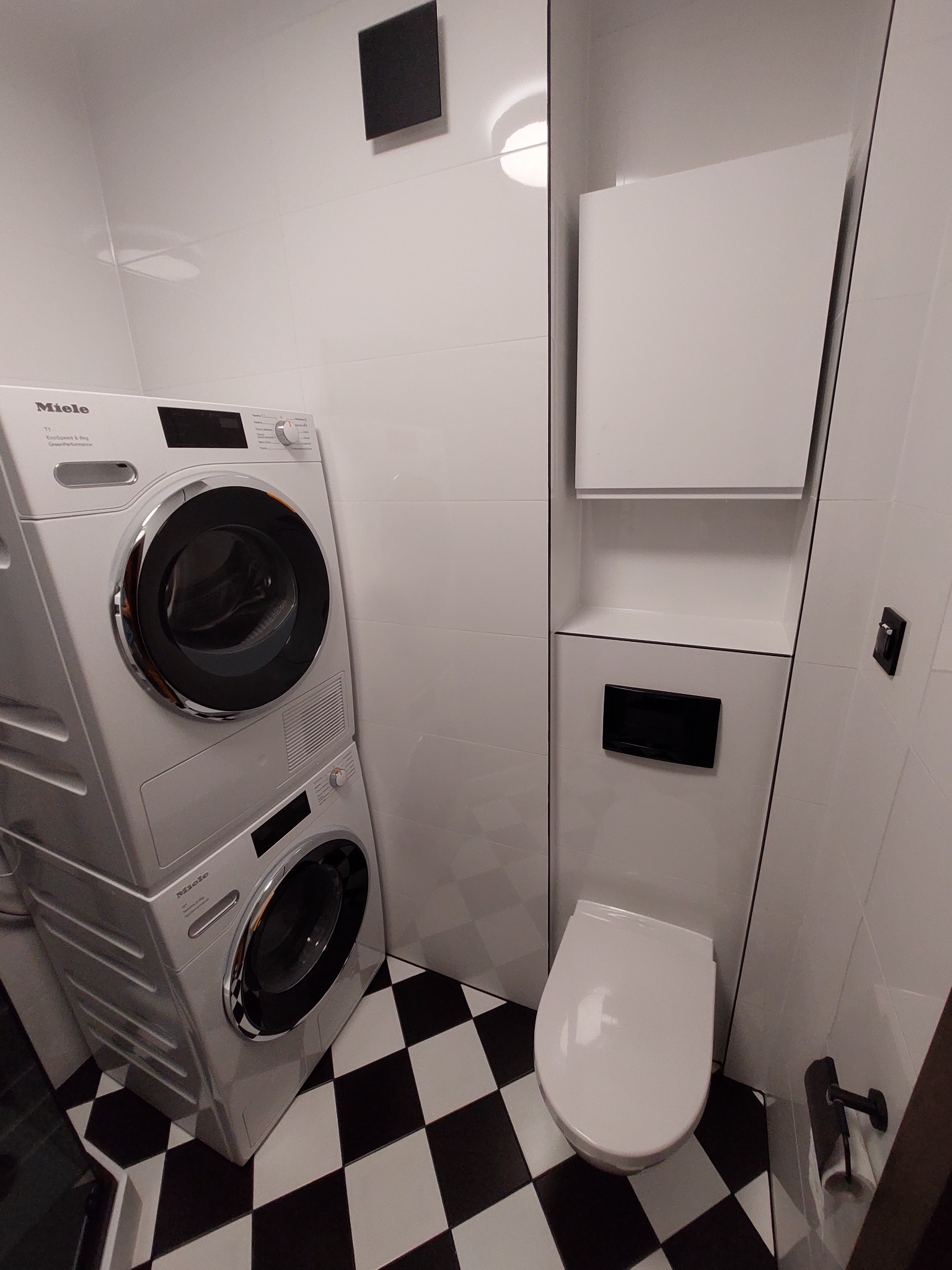 Nowoczesna łazienka z pralką i suszarką Miele ustawionymi w pionie, białą zabudową i toaletą na tle czarno-białej podłogi w szachownicę.